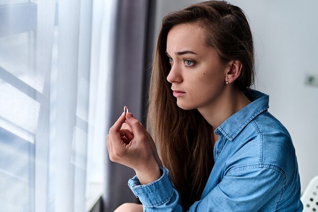 Jovem chateada mulher pensativa solitária deprimida com olhos tristes detém o anel de ouro e senta-se sozinha em casa durante a preocupação com o casamento fracassado após terminar o relacionamento e o divórcio