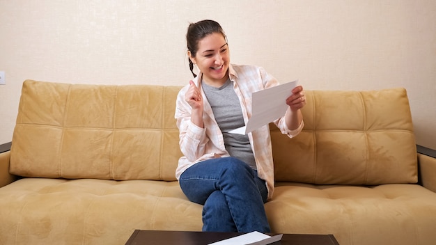 Jovem, chateada, tira o documento de papel do envelope e fica feliz com as boas notícias, sentada em um confortável sofá bege em uma sala de estar iluminada