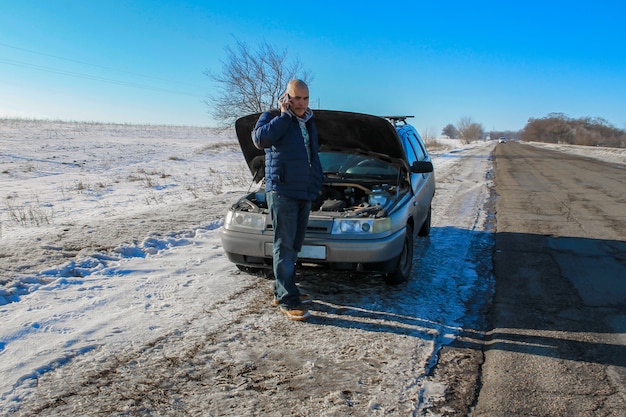 Jovem chateado ligando para o serviço de automóveis perto de um carro quebrado na estrada de inverno