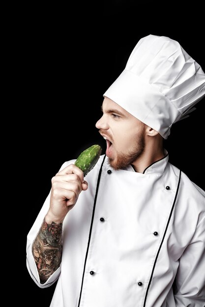 Foto jovem chef barbudo de uniforme branco segurando pepino em fundo preto