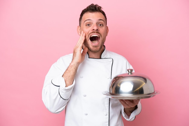 Jovem chef brasileiro com bandeja isolada em fundo rosa com surpresa e expressão facial chocada