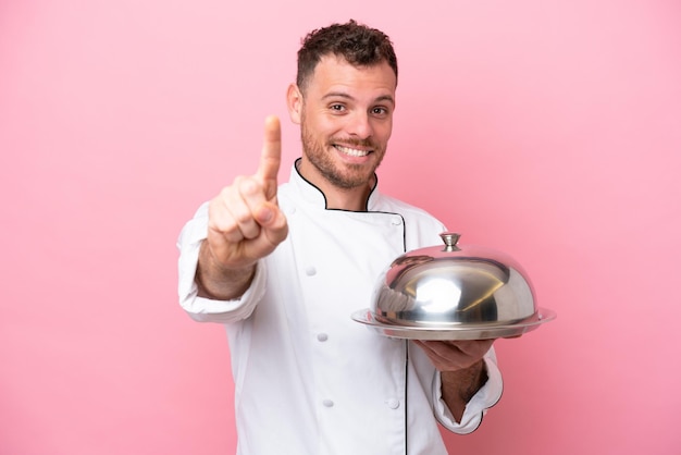 Jovem chef brasileiro com bandeja isolada em fundo rosa mostrando e levantando um dedo