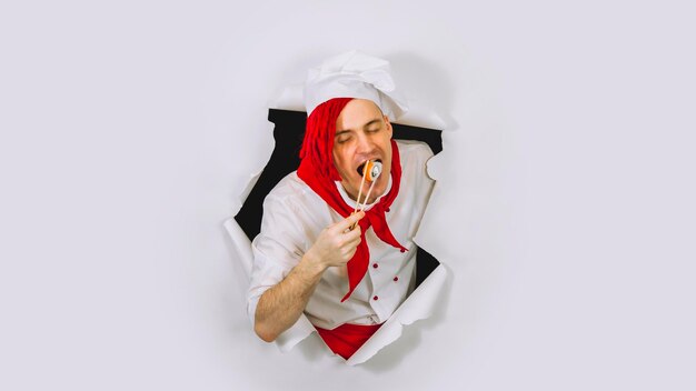 Jovem chef comendo sushi roll no estúdio Cozinheiro masculino de chapéu branco e camisa segura pauzinhos com rolo e come no buraco de fundo branco
