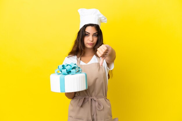 Jovem chef de pastelaria caucasiana com um grande bolo isolado em um fundo amarelo, mostrando o polegar para baixo com expressão negativa