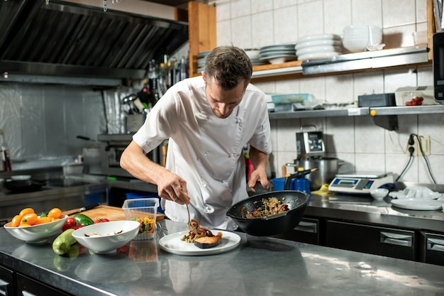 Jovem chef de uniforme segurando uma frigideira com ensopado de vegetais enquanto se inclina sobre a mesa com o prato e coloca a comida em um pedaço de salmão frito