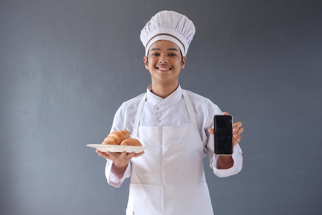 Jovem chef masculino ou padeiro de avental segura um prato de croissant enquanto mostra a tela do smartphone em branco