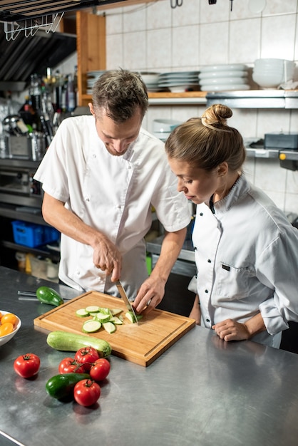 Jovem chef profissional do sexo masculino mostrando a sua estagiária como preparar abobrinhas frescas enquanto os dois estão de pé junto à mesa na cozinha do restaurante