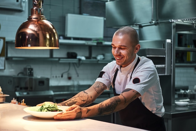 jovem chef sorridente com tatuagens em avental preto segurando um prato pronto na cozinha de um restaurante moderno