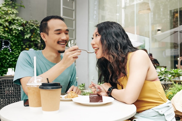 Jovem chinês feliz alimentando a namorada com um pedaço de bolo quando os dois estão sentados à mesa do café