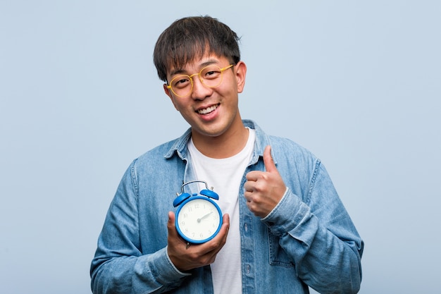 Jovem chinês segurando um despertador, sorrindo e levantando o polegar