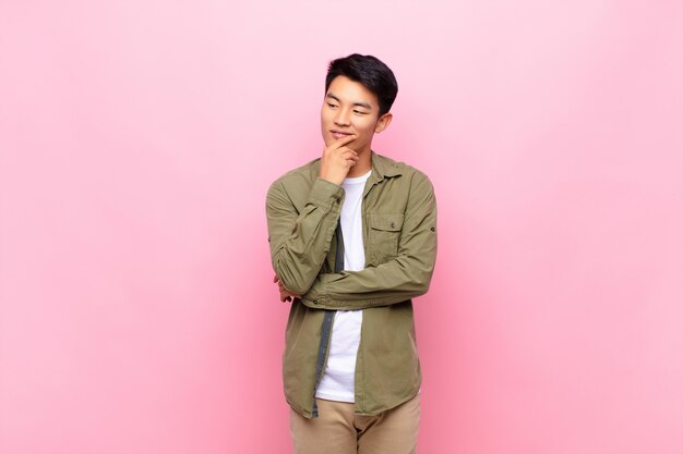 Jovem chinês sorrindo com uma expressão feliz e confiante com a mão no queixo, pensando e olhando para o lado contra uma parede plana de cor