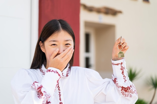 Jovem chinesa ao ar livre segurando as chaves de casa com expressão feliz