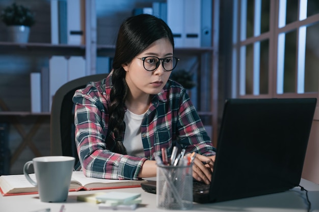 Jovem chinesa asiática adolescente em seu quarto escuro usando computador portátil e conectando-se à internet. estudante universitário concentrado trabalhando duro no notebook pc fazendo projeto com livro.