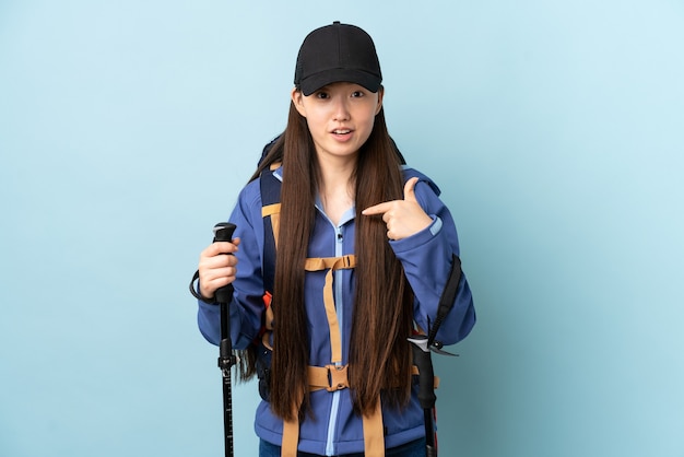 Jovem chinesa com mochila e bastões de trekking na parede azul isolada com expressão facial surpresa