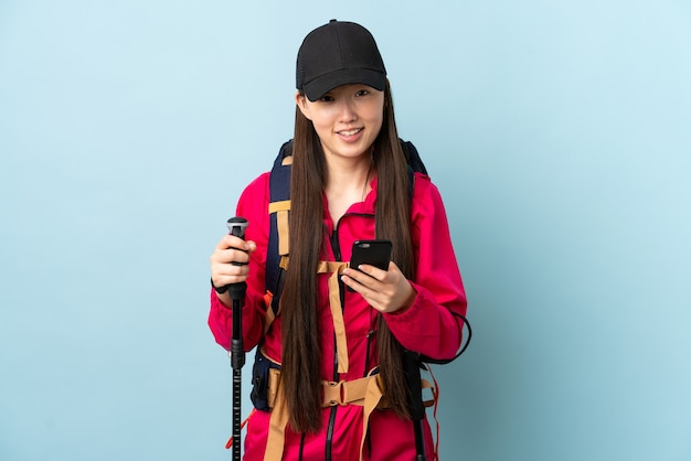 Jovem chinesa com mochila e bastões de trekking sobre azul isolado enviando uma mensagem com o celular
