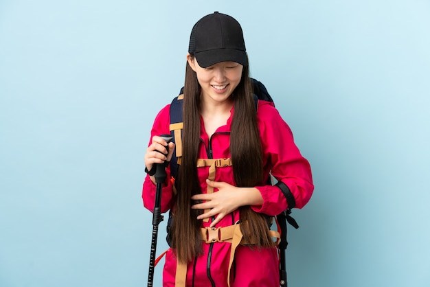 Jovem chinesa com polos mochila e trekking sobre parede azul isolada, sorrindo muito