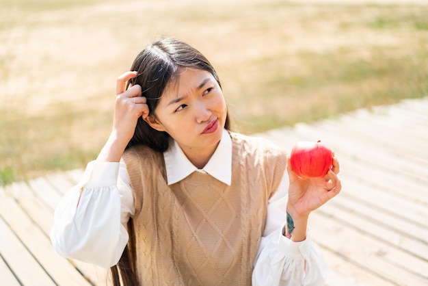 Jovem chinesa com uma maçã ao ar livre com dúvidas e com expressão facial confusa