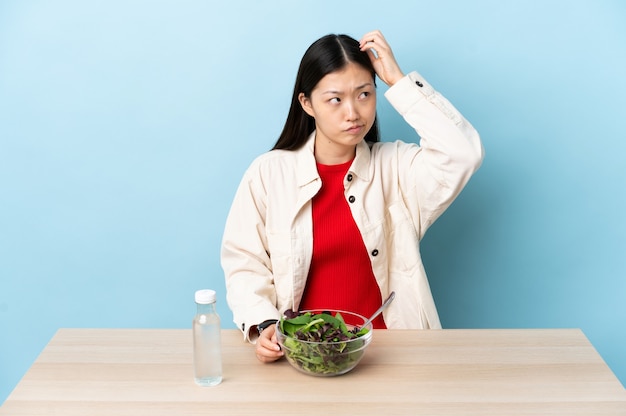 Jovem chinesa comendo uma salada com dúvidas enquanto coça a cabeça