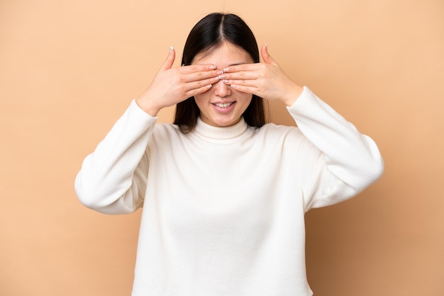 Jovem chinesa isolada em fundo bege, cobrindo os olhos com as mãos e sorrindo