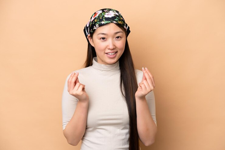 Foto jovem chinesa isolada em fundo bege fazendo gesto de dinheiro