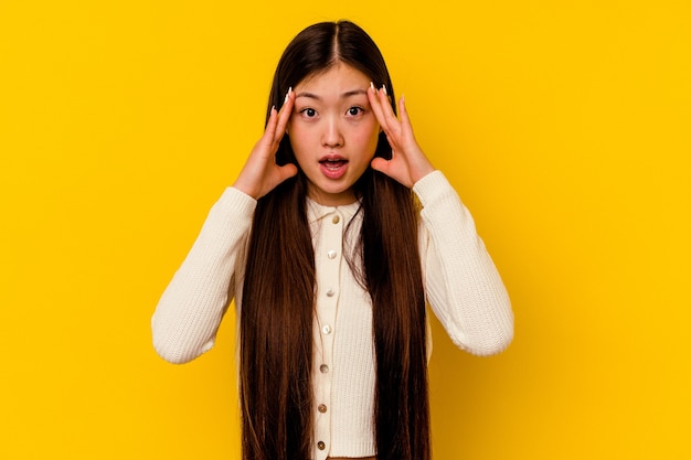 Jovem chinesa isolada em uma parede amarela se divertindo cobrindo metade do rosto com a palma da mão