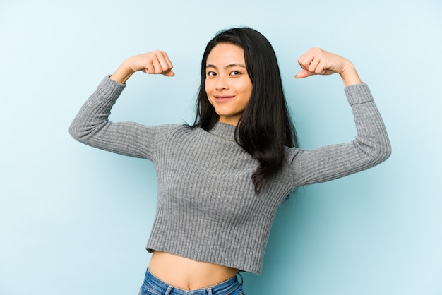 Jovem chinesa isolada em uma parede azul, mostrando o gesto de força com os braços, símbolo do poder feminino