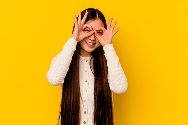 Jovem chinesa isolada na parede amarela mostrando sinal de aprovação sobre os olhos