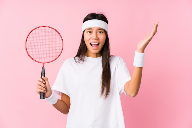 Jovem chinesa jogando badminton em um fundo rosa, recebendo uma surpresa agradável, animado e levantando as mãos.