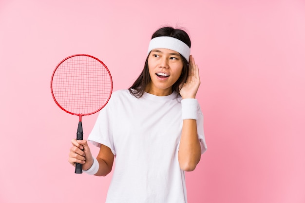 Jovem chinesa jogando badminton em uma parede rosa, tentando ouvir uma fofoca.