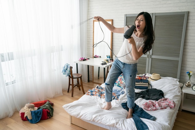 Jovem chinesa muito asiática cantando e dançando com secador de cabelo em vez de microfone em pé na cama como super estrela no palco em concerto. adolescente fazendo mala no quarto em casa