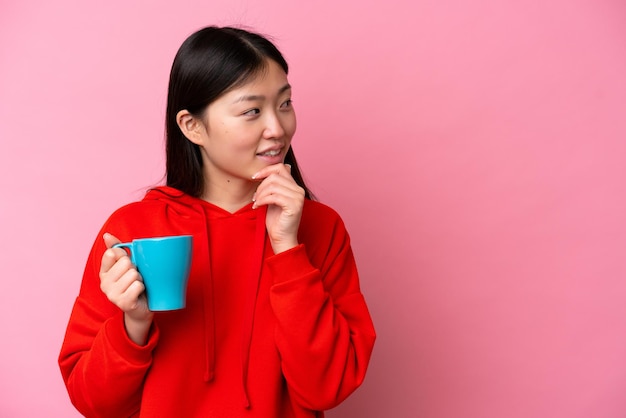 Jovem chinesa segurando a xícara de café isolada no fundo rosa pensando em uma ideia e olhando de lado