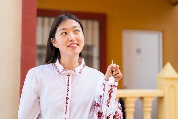 Jovem chinesa segurando as chaves de casa ao ar livre olhando para cima enquanto sorri