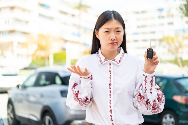 Jovem chinesa segurando as chaves do carro ao ar livre fazendo gesto de dúvidas enquanto levanta os ombros