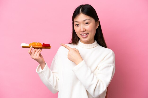 Jovem chinesa segurando sashimi isolado em fundo rosa apontando para o lado para apresentar um produto