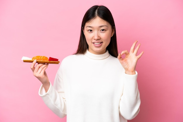 Jovem chinesa segurando sashimi isolado em fundo rosa mostrando sinal de ok com os dedos