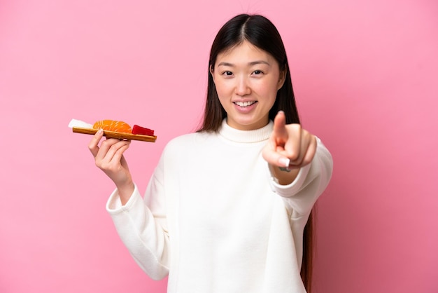 Jovem chinesa segurando sashimi isolado no fundo rosa apontando para a frente com expressão feliz