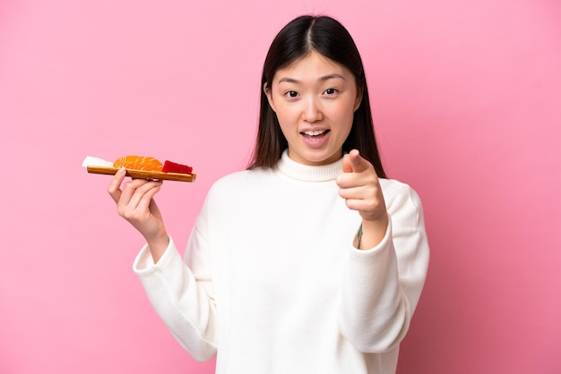 Jovem chinesa segurando sashimi isolado no fundo rosa surpreso e apontando para a frente