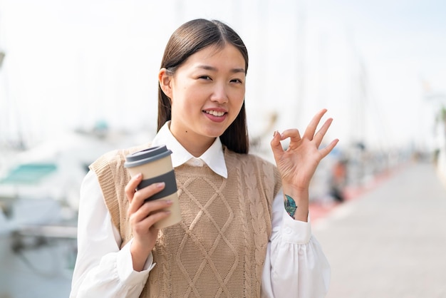 Jovem chinesa segurando um café para viagem ao ar livre mostrando sinal de ok com os dedos