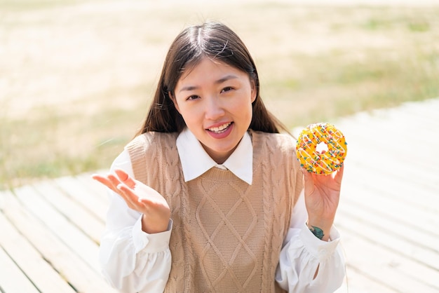 Jovem chinesa segurando um donut ao ar livre com expressão facial chocada