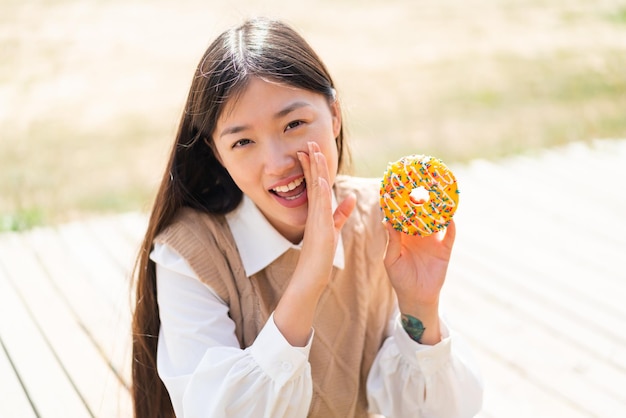 Jovem chinesa segurando um donut ao ar livre sussurrando algo