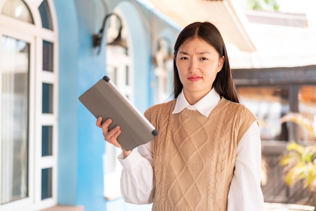 Jovem chinesa segurando um tablet ao ar livre com expressão triste