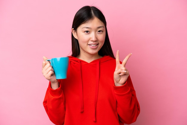 Jovem chinesa segurando xícara de café isolada em fundo rosa sorrindo e mostrando sinal de vitória