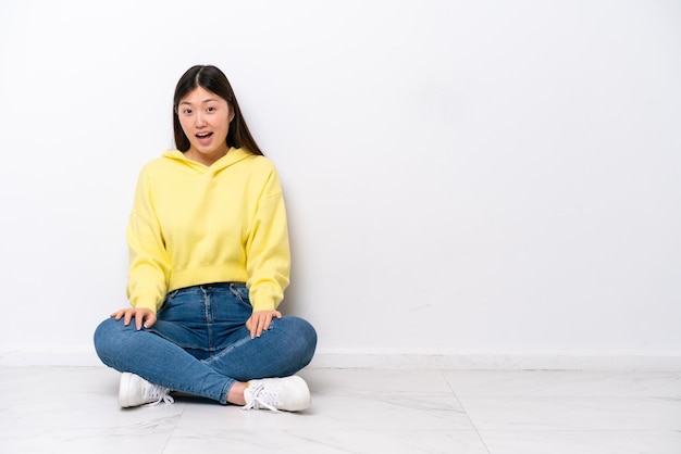 Jovem chinesa sentada no chão isolada na parede branca com expressão facial de surpresa