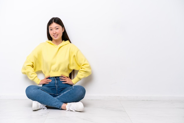 Jovem chinesa sentada no chão isolada na parede branca posando com os braços no quadril e sorrindo