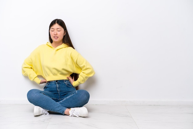 Jovem chinesa sentada no chão isolada na parede branca sofrendo de dor nas costas por ter feito um esforço