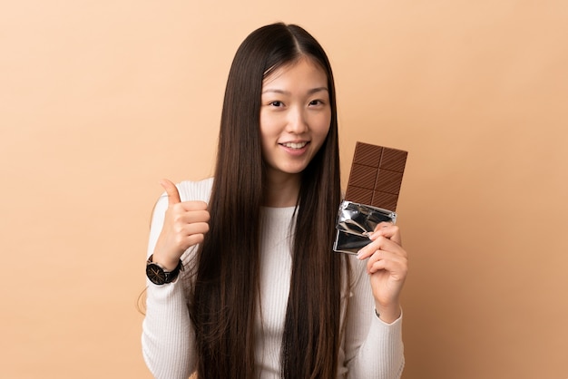 Jovem chinesa sobre parede isolada, tomando um tablete de chocolate e com o polegar