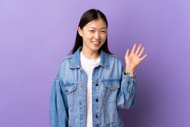 Jovem chinesa sobre parede roxa isolada saudando com mão com expressão feliz