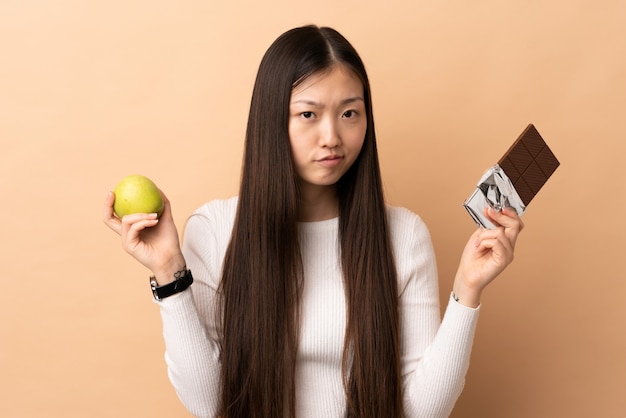 Jovem chinesa tendo dúvidas enquanto toma uma tablete de chocolate em uma mão e uma maçã na outra