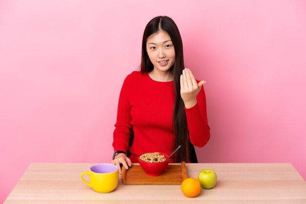 Jovem chinesa tomando café da manhã em uma mesa convidativa para vir com a mão. feliz que você veio