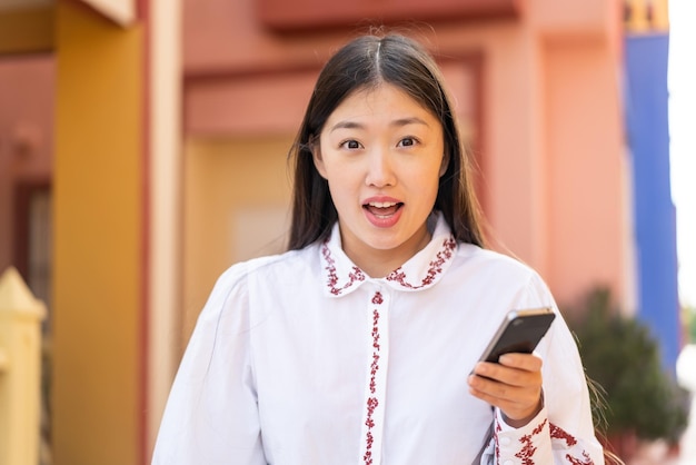 Jovem chinesa usando telefone celular ao ar livre com surpresa e expressão facial chocada